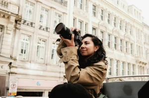 Woman in trenchcoat taking a photo in front of a building with three rows of windows